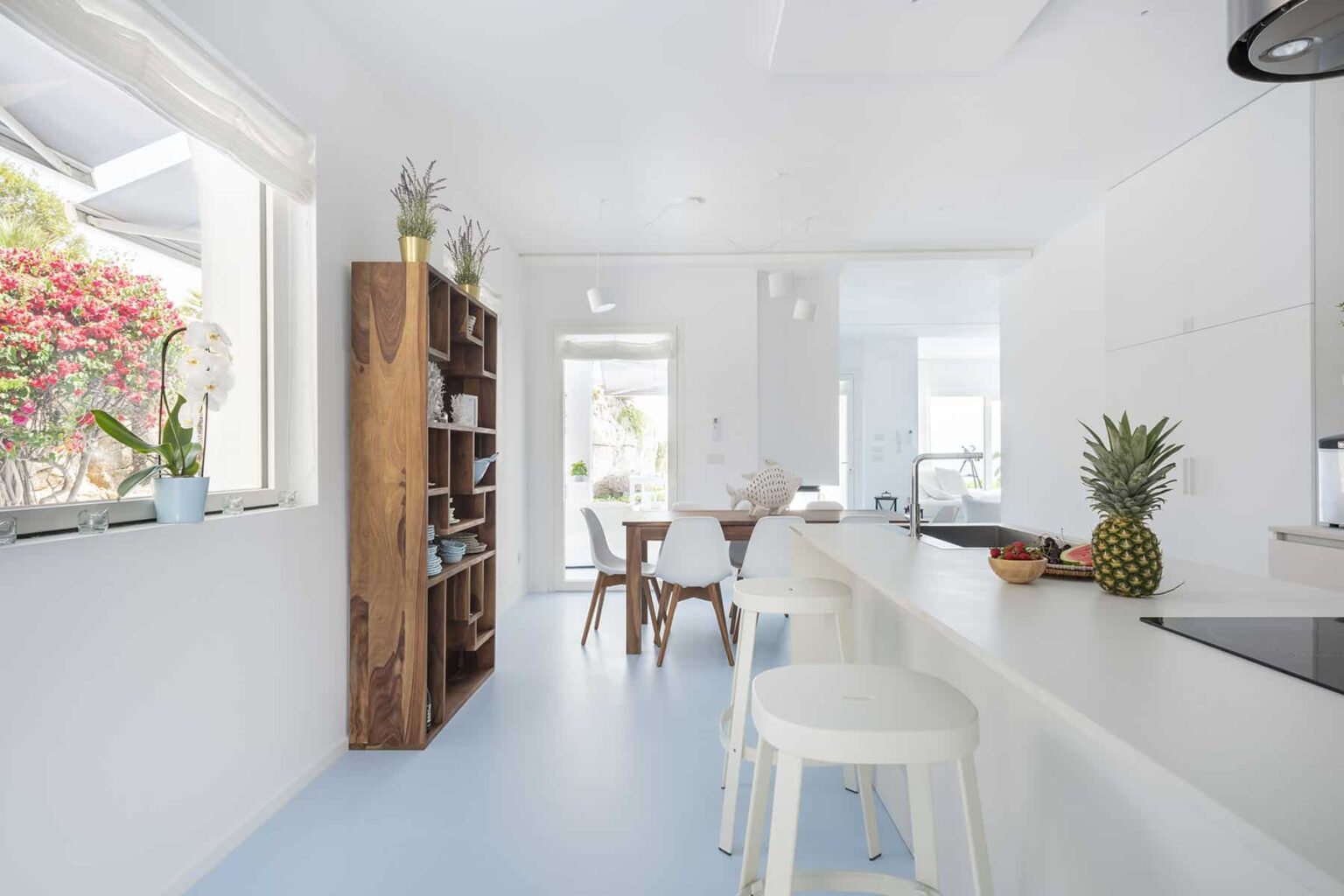 Modern bright kitchen featuring white cabinetry, a wooden bookshelf, and a dining area with a wooden table and chairs. Natural light floods the space, complemented by indoor plants and a pineapple on the counter. | Only on SopranoVillas