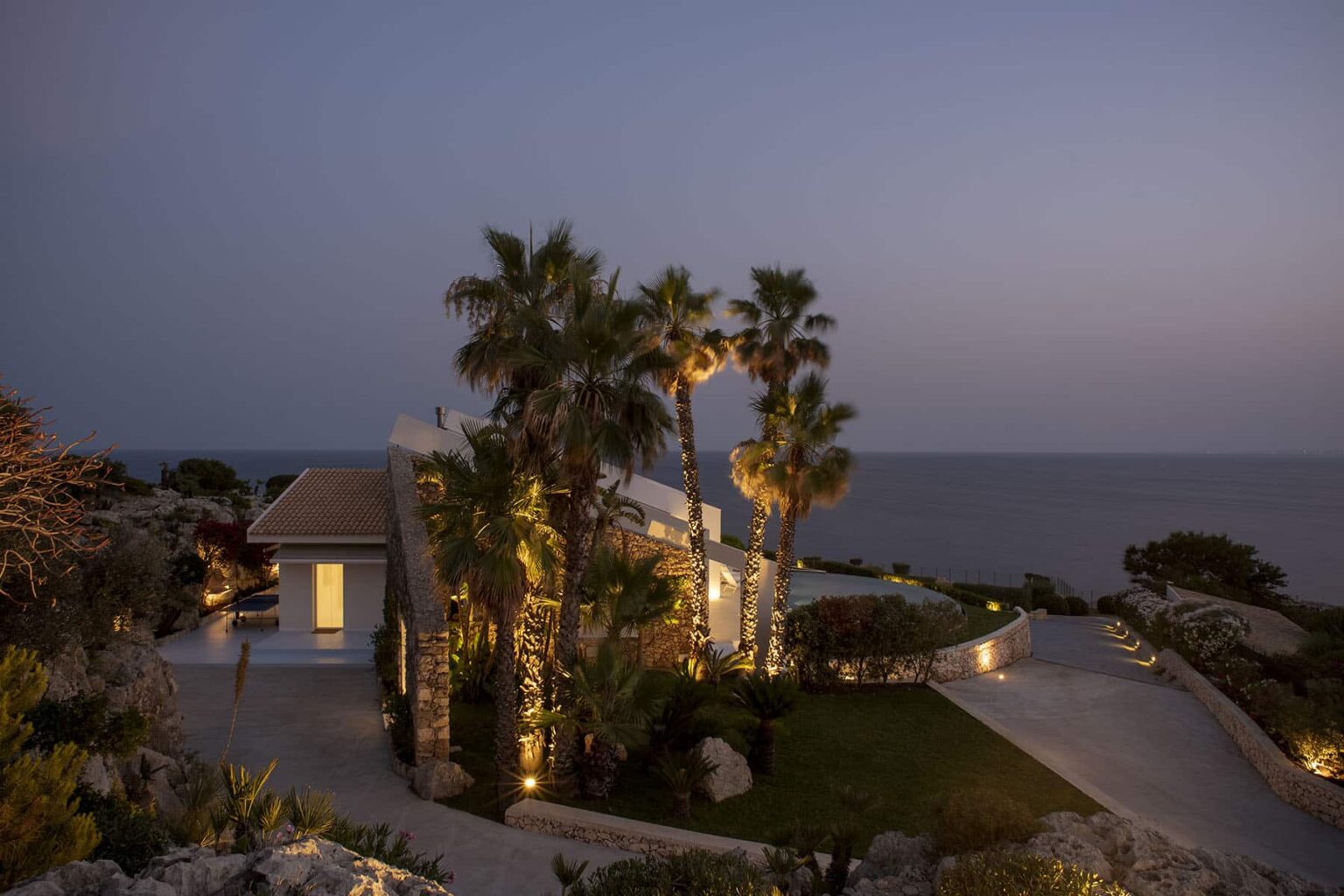 Coastal villa at dusk with palm trees and ocean view, featuring warm outdoor lighting and a welcoming pathway. Perfect for luxury vacation rentals or seaside retreats. | Only on SopranoVillas