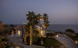 Coastal villa at dusk with palm trees and ocean view, featuring warm outdoor lighting and a welcoming pathway. Perfect for luxury vacation rentals or seaside retreats.