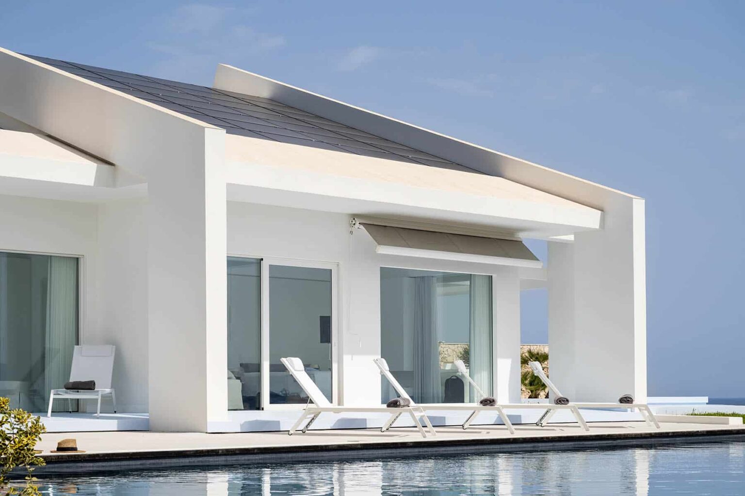 Modern white villa with large windows, sun loungers by the pool, and a clear blue sky in the background. Ideal for luxury vacation rentals or contemporary architecture inspiration. | Only on SopranoVillas