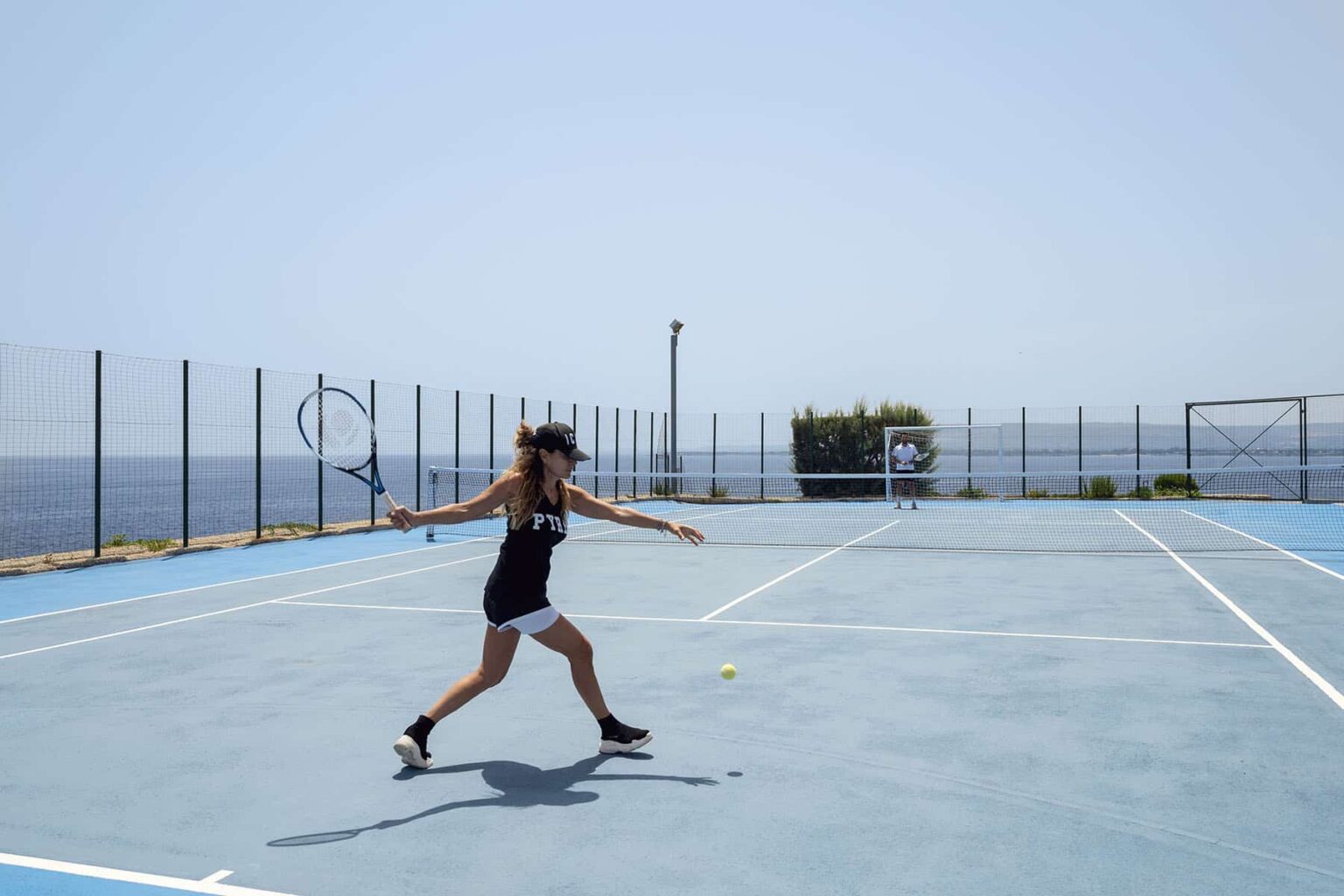 Woman playing tennis on a blue court by the sea, demonstrating an athletic stance and swing under clear skies. | Only on SopranoVillas