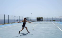Woman playing tennis on a blue court by the sea, demonstrating an athletic stance and swing under clear skies.