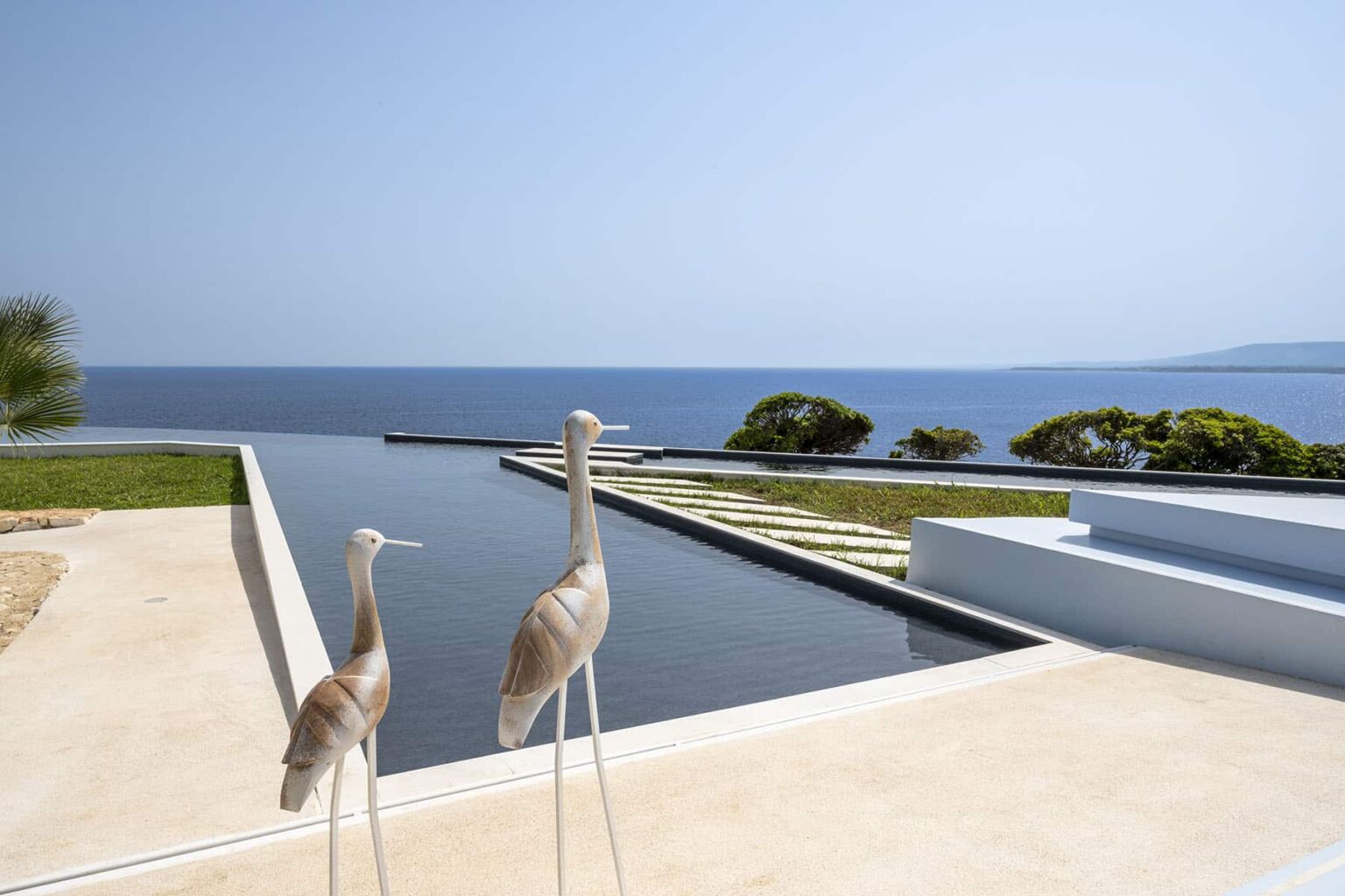 Two elegant sculptures of herons stand by a serene infinity pool overlooking the ocean, with a clear blue sky and lush greenery in the background, showcasing a tranquil coastal landscape. | Only on SopranoVillas