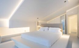 Modern minimalist bedroom featuring a large white bed with crisp linens, soft ambient lighting, and a sleek design. The room emphasizes clean lines and a spacious feel, ideal for relaxation and comfort.