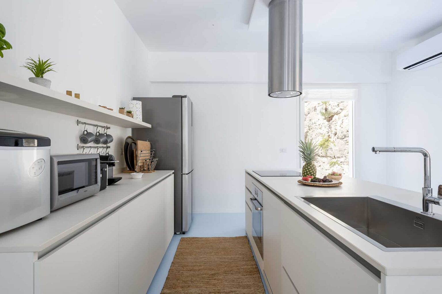 Modern kitchen interior featuring sleek white cabinetry, stainless steel appliances, and a minimalist design. The space includes a double sink, a stylish countertop, and natural light streaming through a window, complemented by decorative plants. | Only on SopranoVillas
