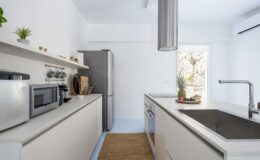 Modern kitchen interior featuring sleek white cabinetry, stainless steel appliances, and a minimalist design. The space includes a double sink, a stylish countertop, and natural light streaming through a window, complemented by decorative plants.