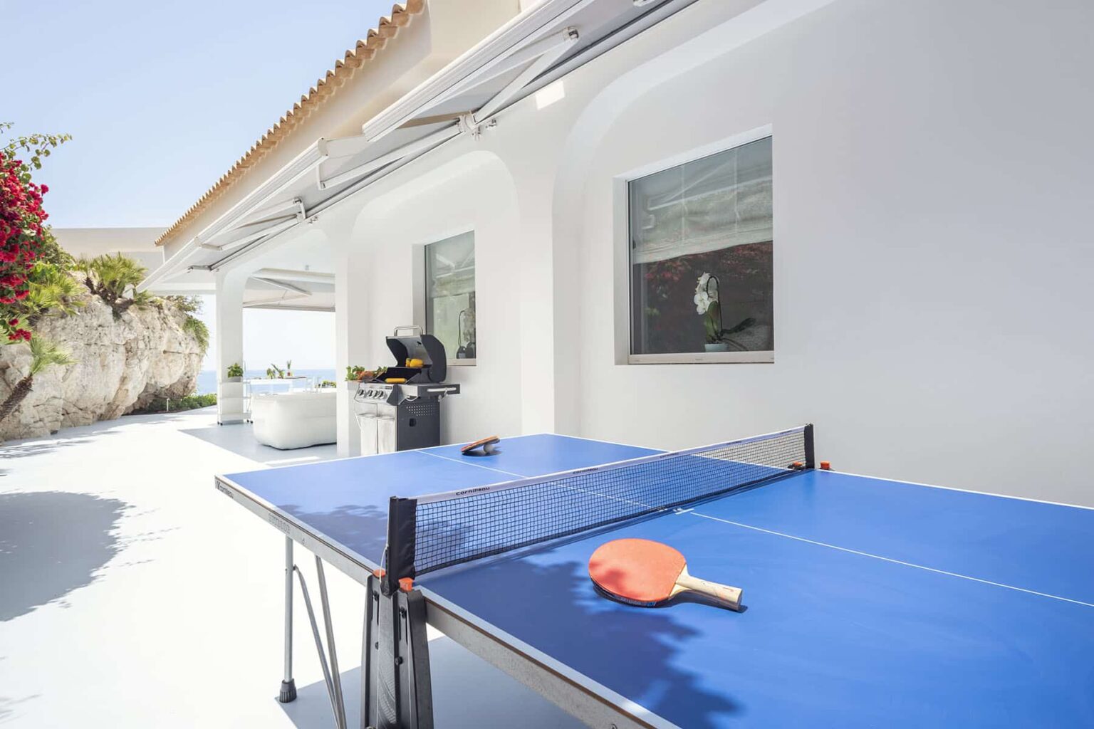 Outdoor ping pong table with a blue surface and an orange paddle, set in a bright, modern patio area with white walls and lush greenery in the background. | Only on SopranoVillas