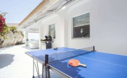 Outdoor ping pong table with a blue surface and an orange paddle, set in a bright, modern patio area with white walls and lush greenery in the background.