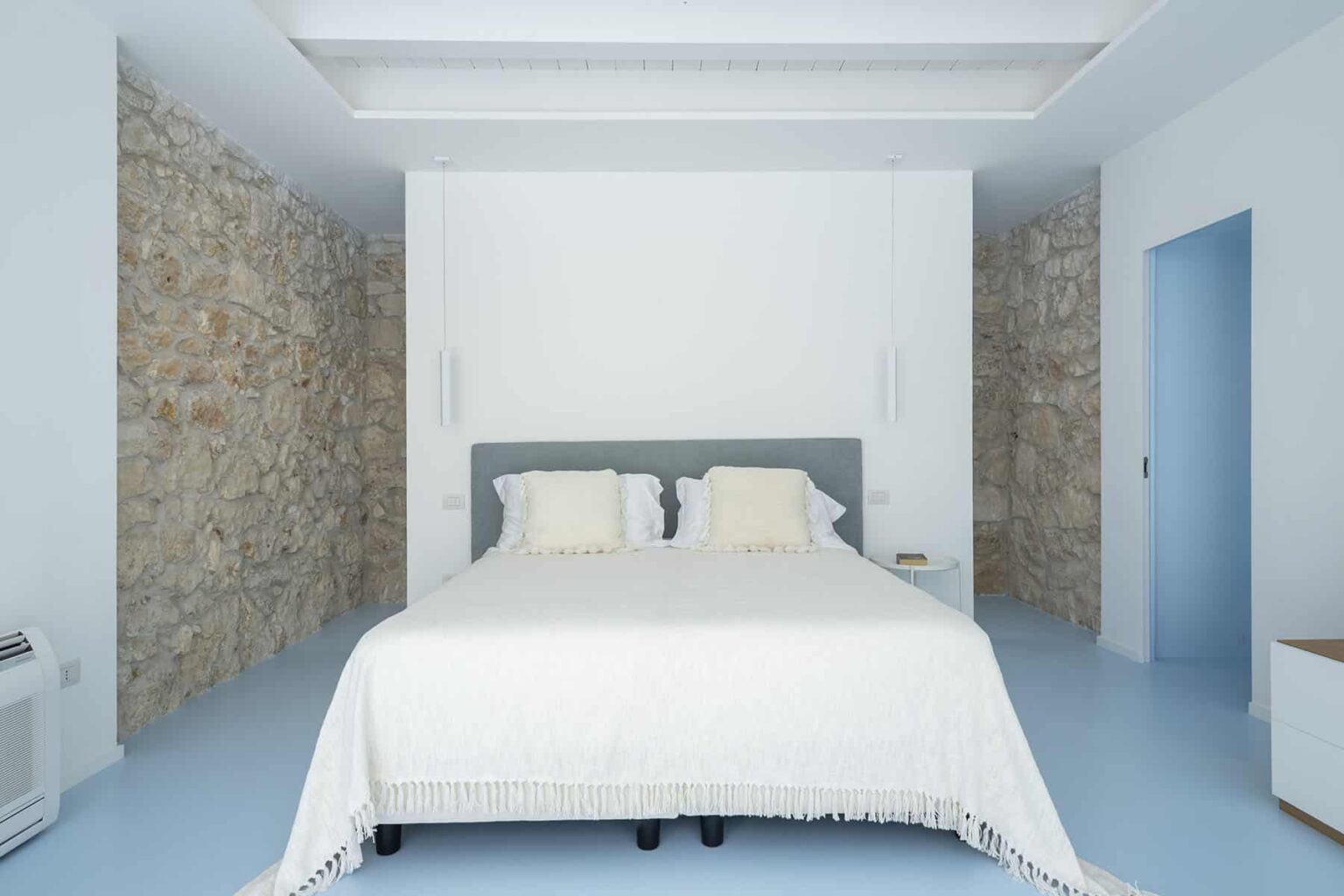 Modern bedroom featuring a spacious double bed with a light-colored blanket, complemented by a minimalist design and stone accent walls. The room has a serene blue floor and a subtle color palette, creating a calming atmosphere. | Only on SopranoVillas