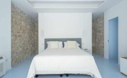 Modern bedroom featuring a spacious double bed with a light-colored blanket, complemented by a minimalist design and stone accent walls. The room has a serene blue floor and a subtle color palette, creating a calming atmosphere.