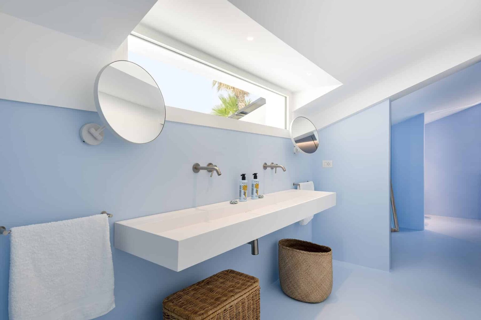 Modern bathroom with a minimalist design featuring a white floating sink, two round mirrors, and a light blue color scheme. Natural light enters through a skylight, enhancing the spacious feel. A woven basket and a towel add functional decor elements. | Only on SopranoVillas