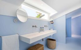 Modern bathroom with a minimalist design featuring a white floating sink, two round mirrors, and a light blue color scheme. Natural light enters through a skylight, enhancing the spacious feel. A woven basket and a towel add functional decor elements.