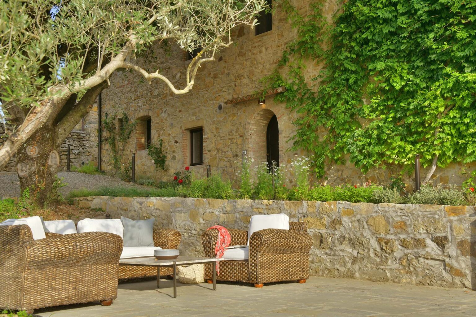 Cozy outdoor seating area with wicker chairs and a coffee table, surrounded by lush greenery and a stone wall, set against a rustic building with climbing vines. Ideal for relaxation and enjoying nature. | Only on SopranoVillas
