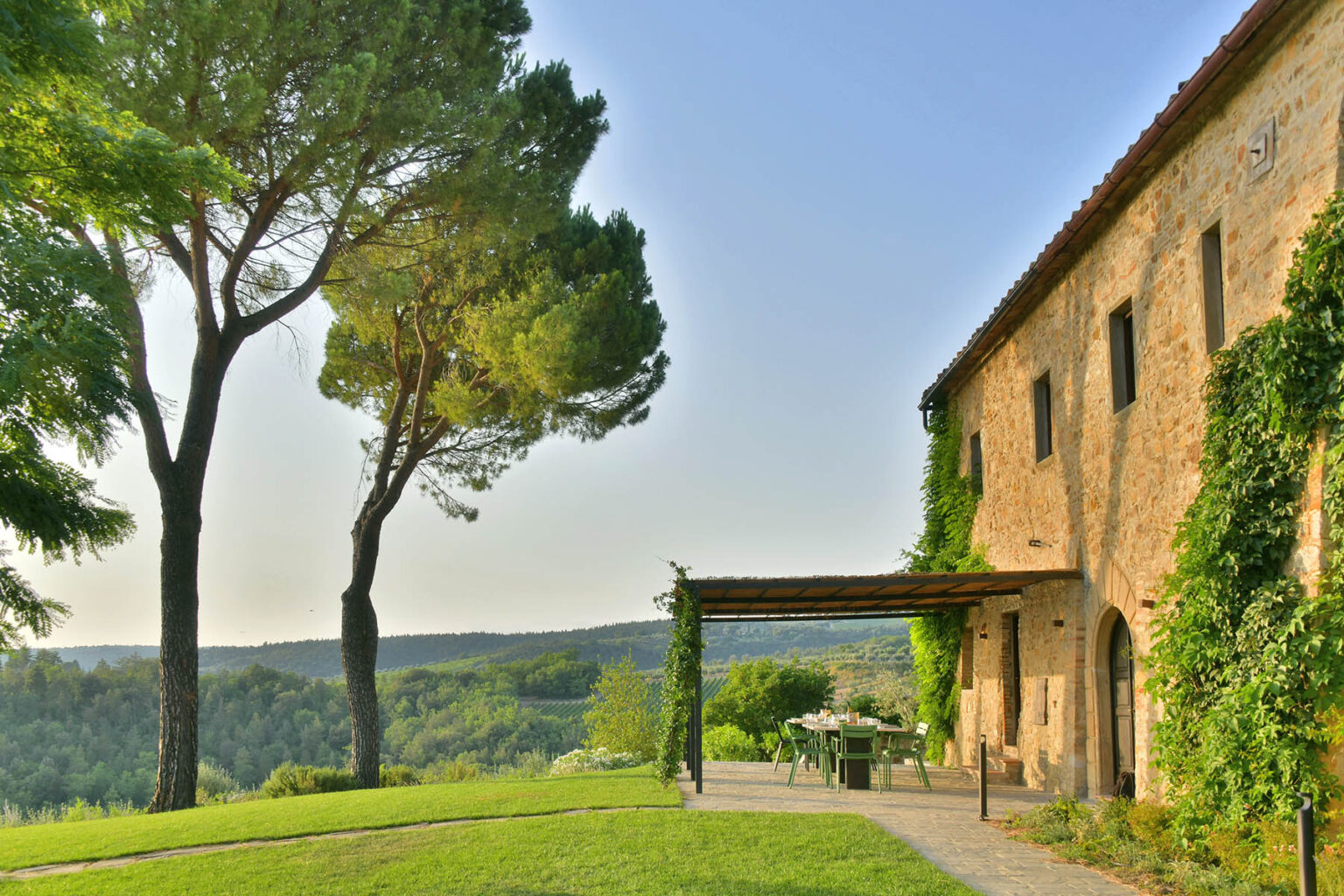 Scenic view of a rustic stone house surrounded by lush greenery and tall trees, with a patio area overlooking rolling hills in the background, ideal for tranquil outdoor settings. | Only on SopranoVillas