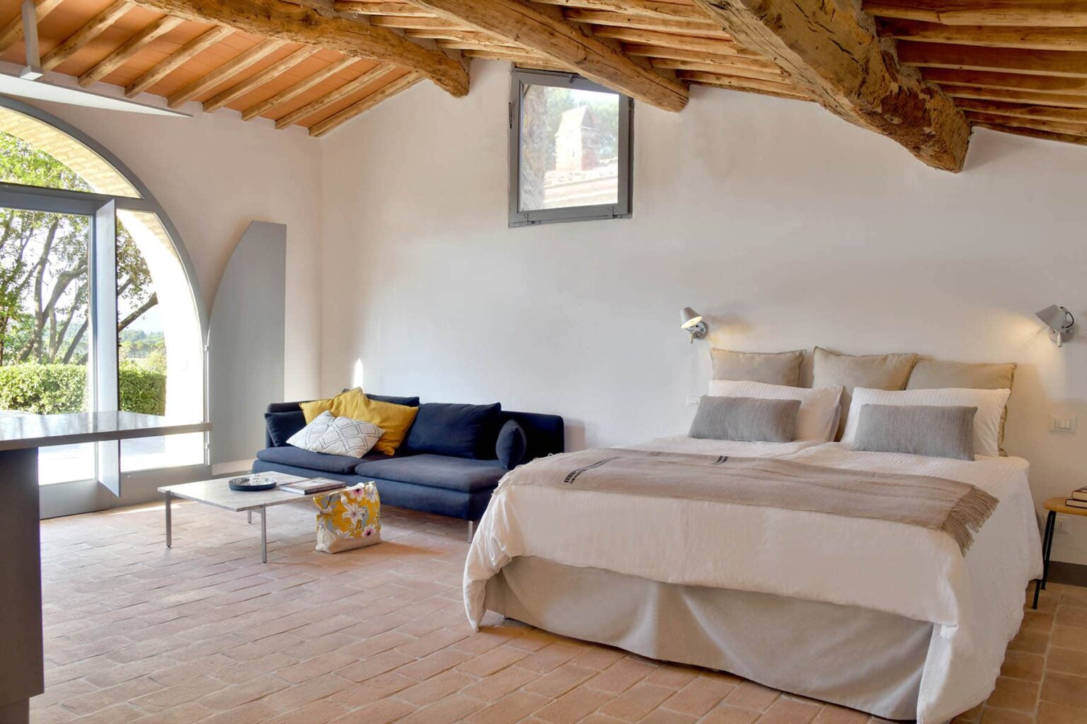 Modern bedroom interior featuring a large bed with neutral bedding, a cozy sofa, wooden ceiling beams, and natural light from arched windows. Ideal for vacation rentals or home decor inspiration. | Only on SopranoVillas