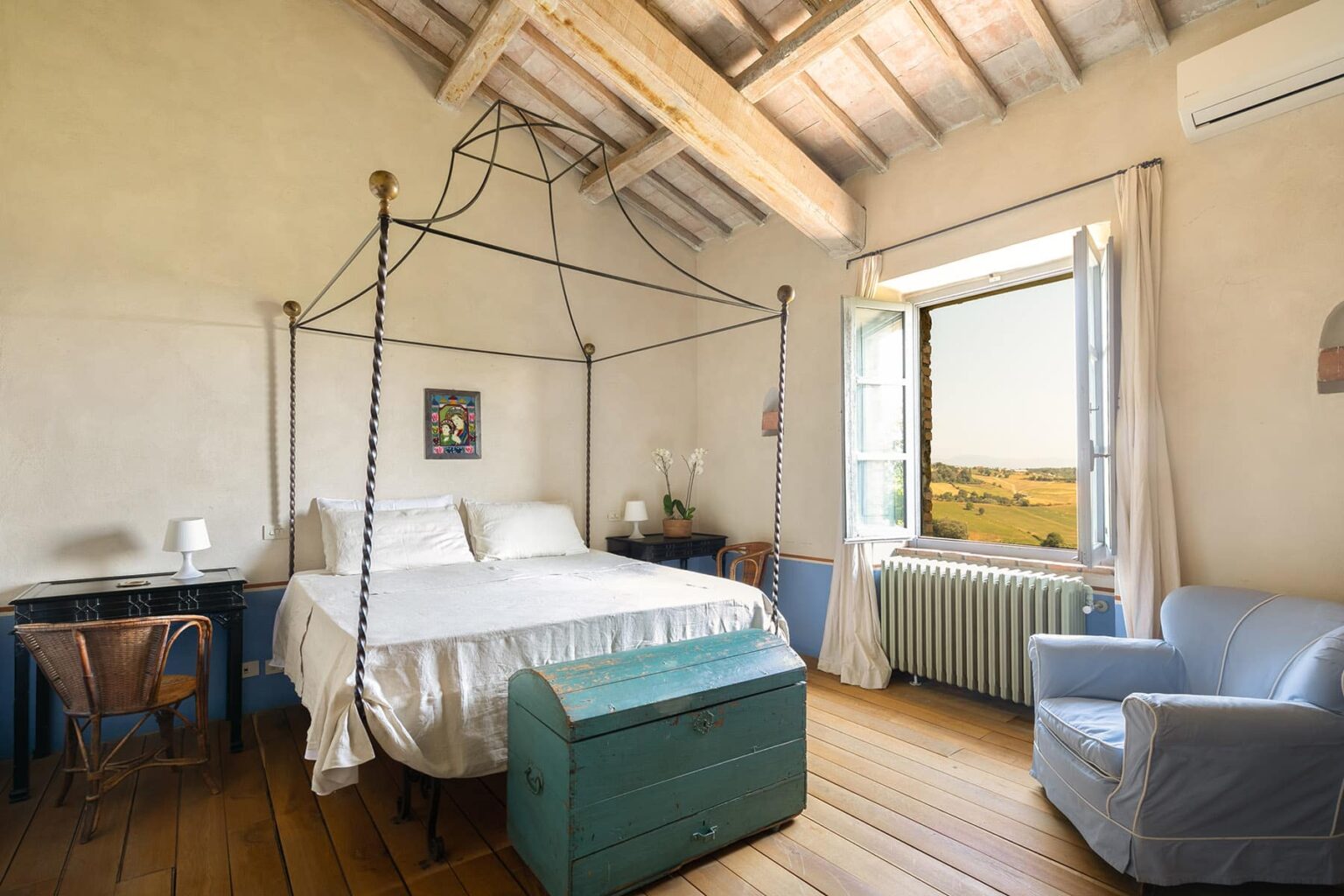 Cozy bedroom featuring a four-poster bed, rustic wooden beams, and a scenic view through a large window. The room includes a bedside table, a vintage blue chest, and a comfortable armchair, creating a warm and inviting atmosphere. | Only on SopranoVillas