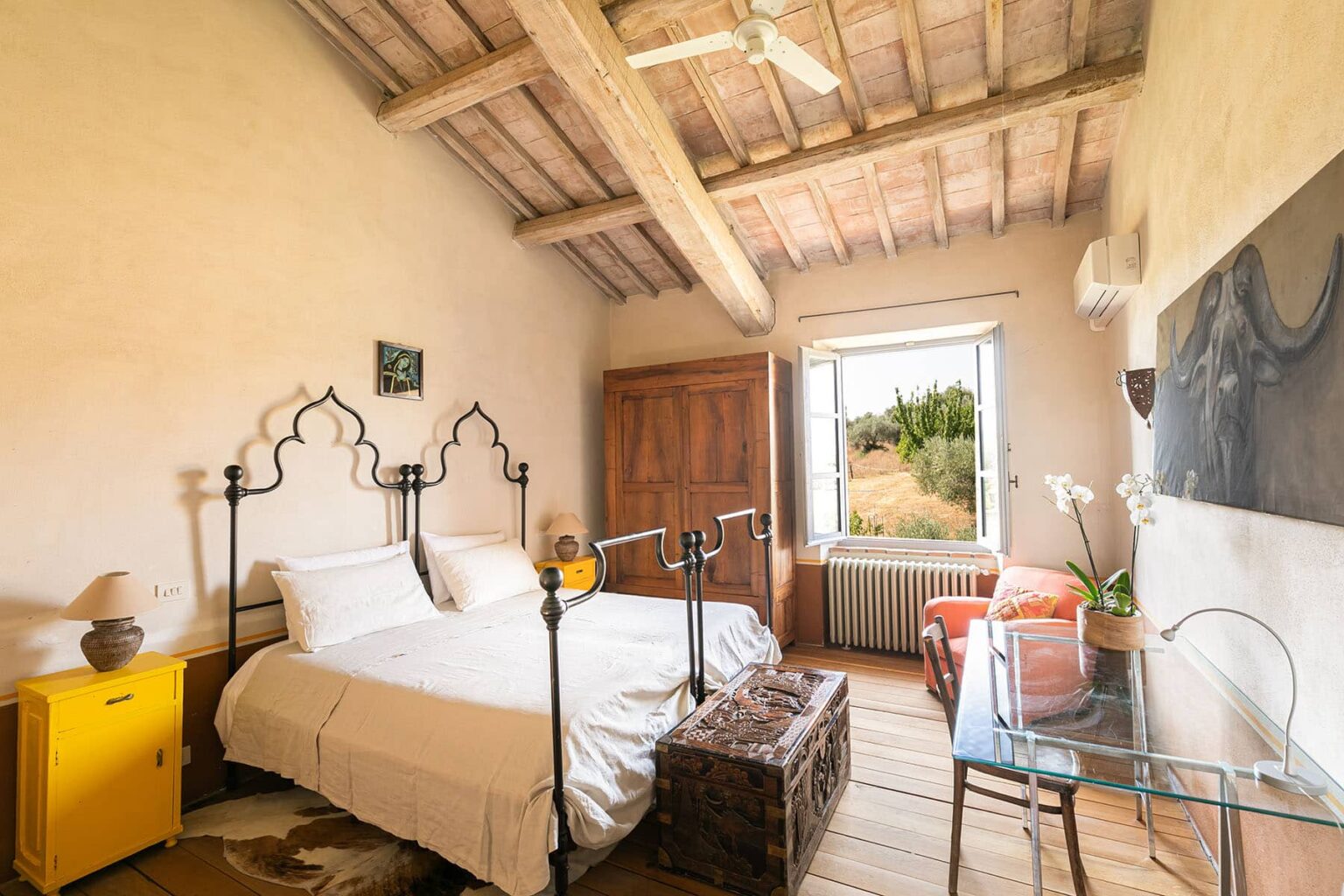 Cozy bedroom featuring a double bed with an ornate metal frame, rustic wooden beams, and a bright window overlooking a scenic view. The room includes a stylish yellow nightstand, a comfortable armchair, and a small decorative table, creating a warm and inviting atmosphere. | Only on SopranoVillas