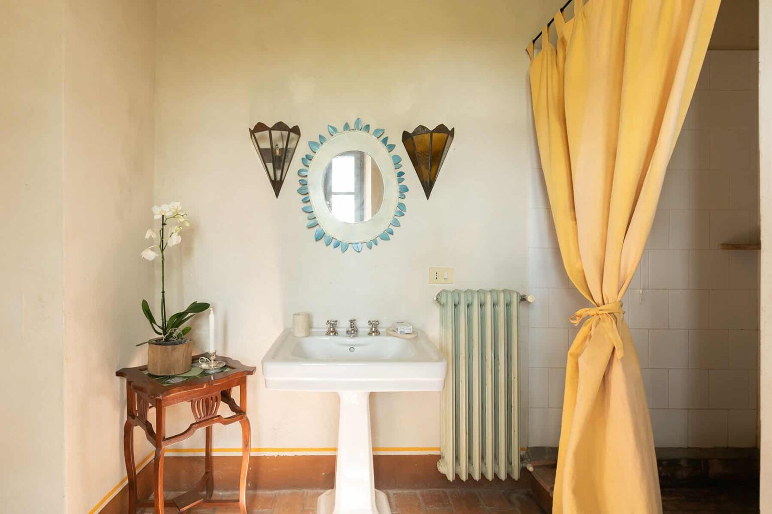 Interior view of a stylish bathroom featuring a white sink, decorative mirror, and a yellow shower curtain, complemented by a vintage wooden table and a potted orchid. The space is bright and inviting, showcasing a blend of modern and classic design elements. | Only on SopranoVillas