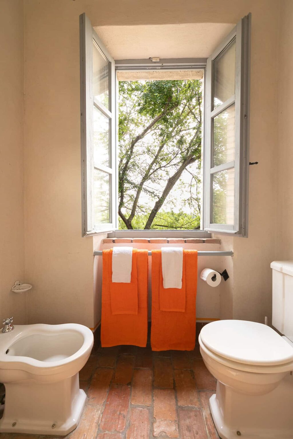 Bright bathroom featuring open windows with a view of greenery, orange towels hanging on a towel rack, and a roll of toilet paper nearby. | Only on SopranoVillas