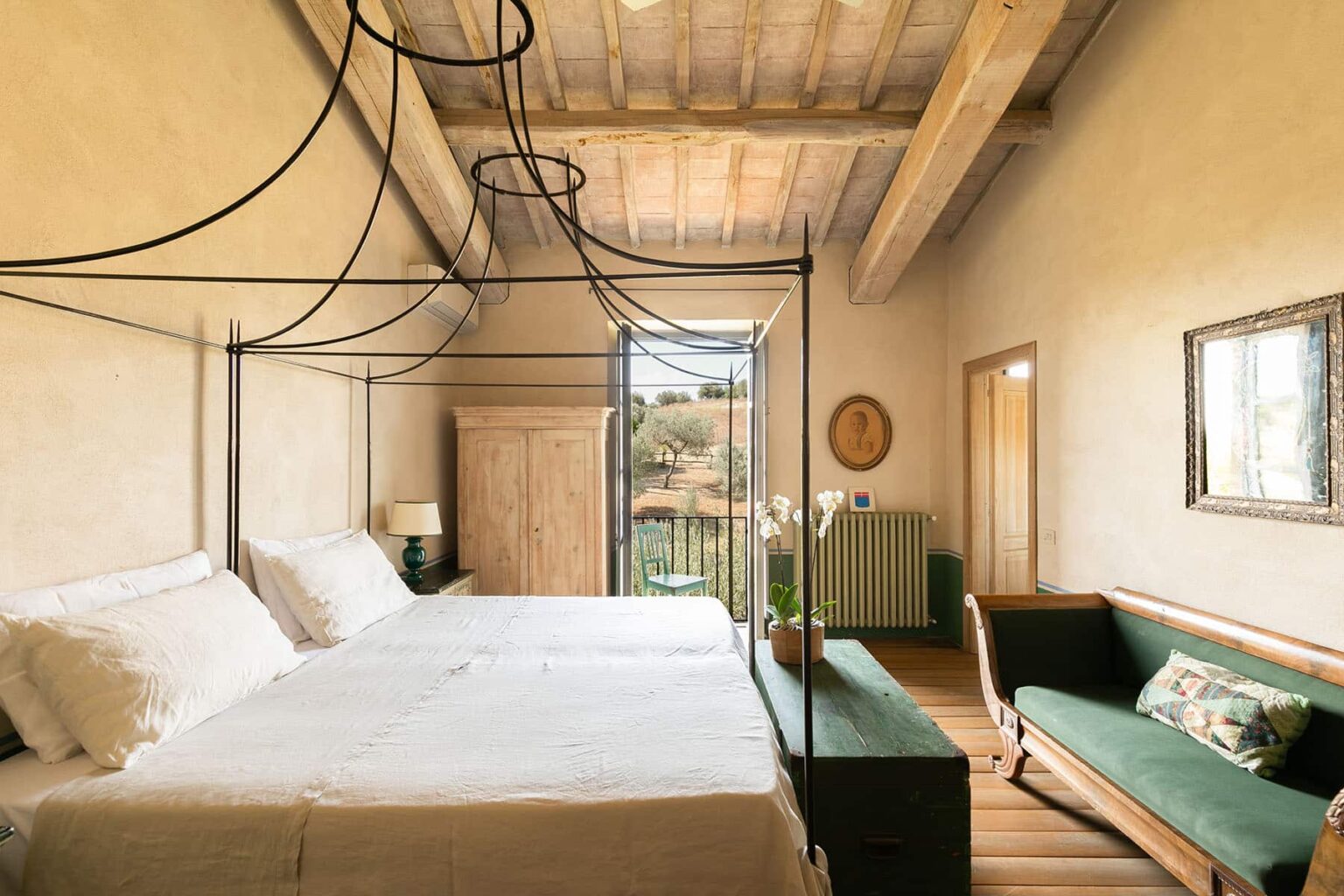 Spacious and elegantly designed bedroom featuring a four-poster bed, wooden beams, and natural light streaming through large windows, complemented by stylish furnishings and a cozy seating area. | Only on SopranoVillas