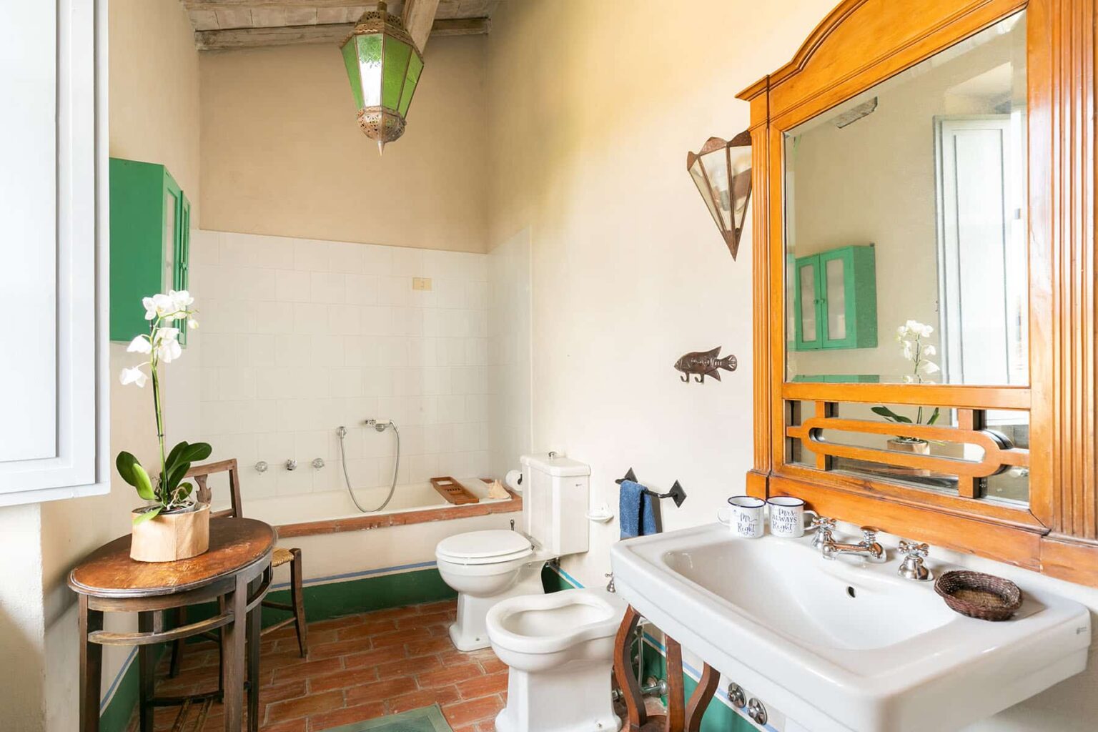 Bright and airy bathroom featuring a wooden vanity with a large mirror, a white sink, a toilet, and a shower area. The space is adorned with green accents, a potted plant, and a rustic wooden table, creating a welcoming atmosphere. | Only on SopranoVillas
