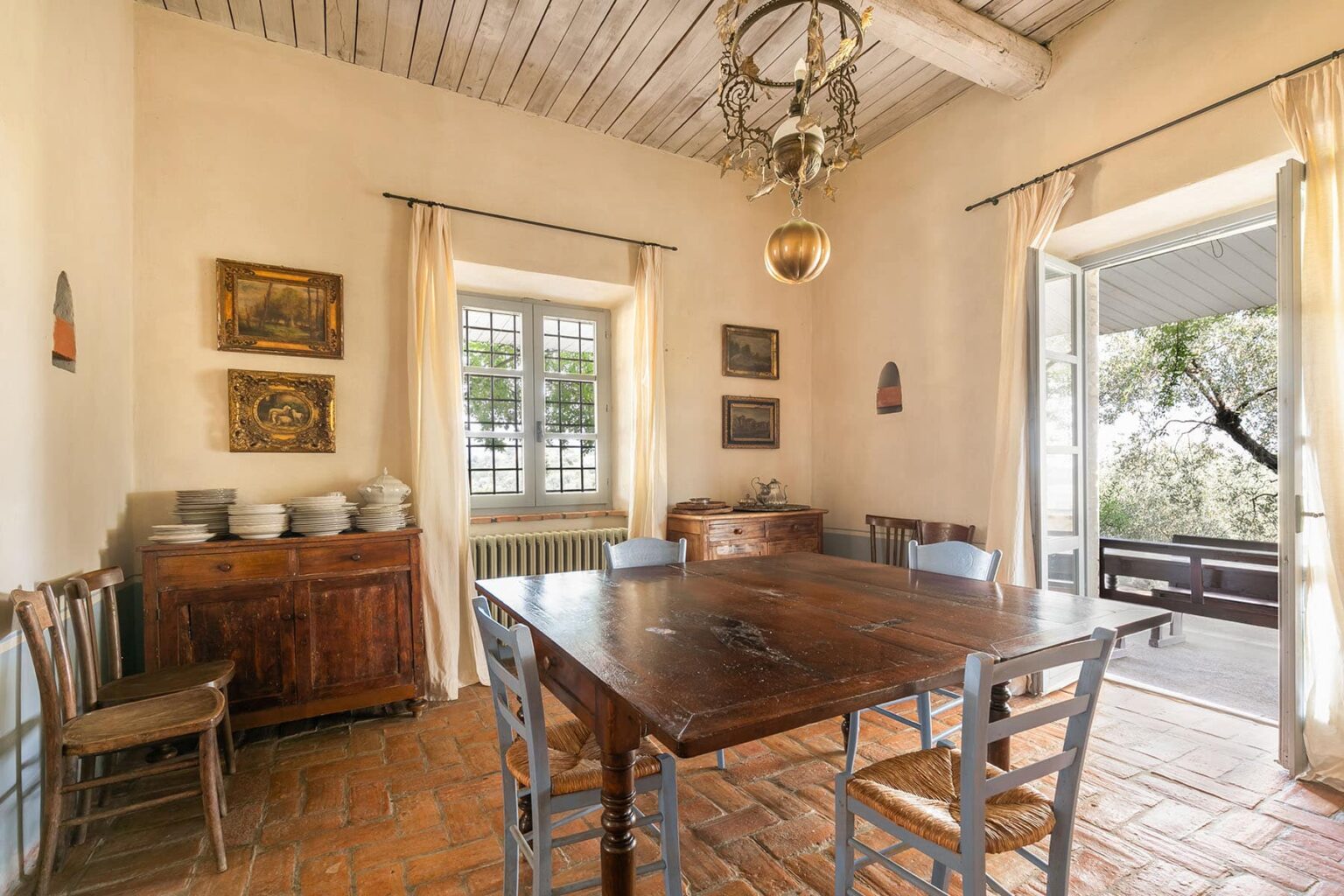 Cozy dining room featuring a wooden table surrounded by chairs, with a rustic chandelier and artwork on the walls, opening to a view of greenery through large windows. | Only on SopranoVillas