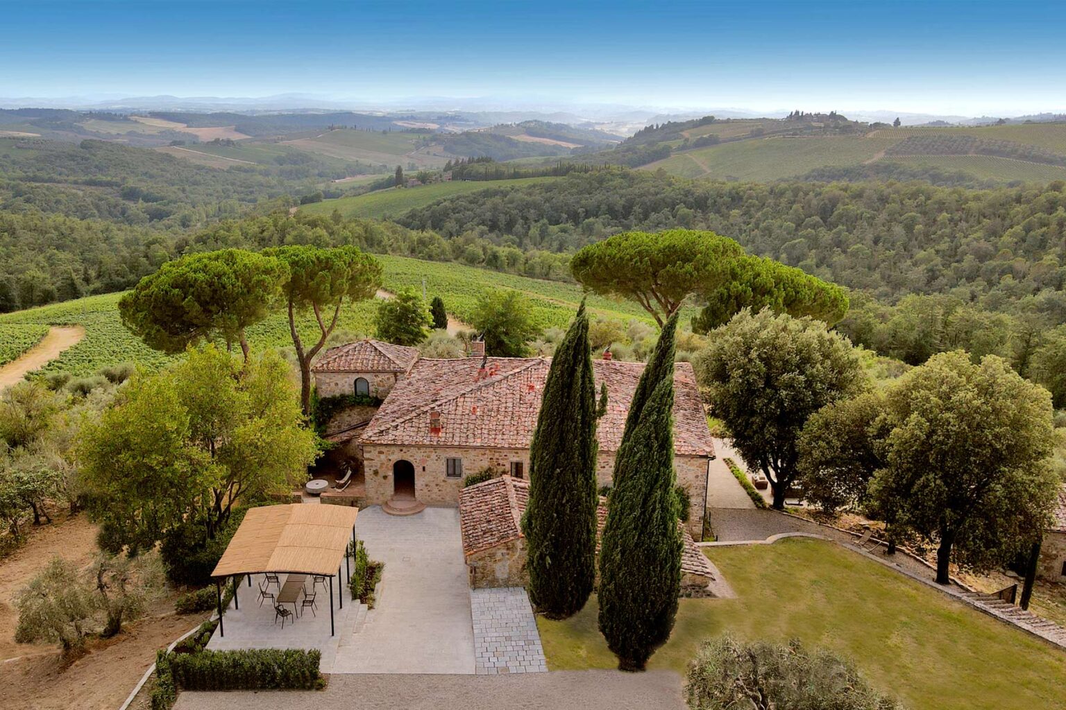Aerial view of a charming rustic villa surrounded by lush green hills and cypress trees in a picturesque landscape, ideal for vacation rentals or countryside retreats. | Only on SopranoVillas