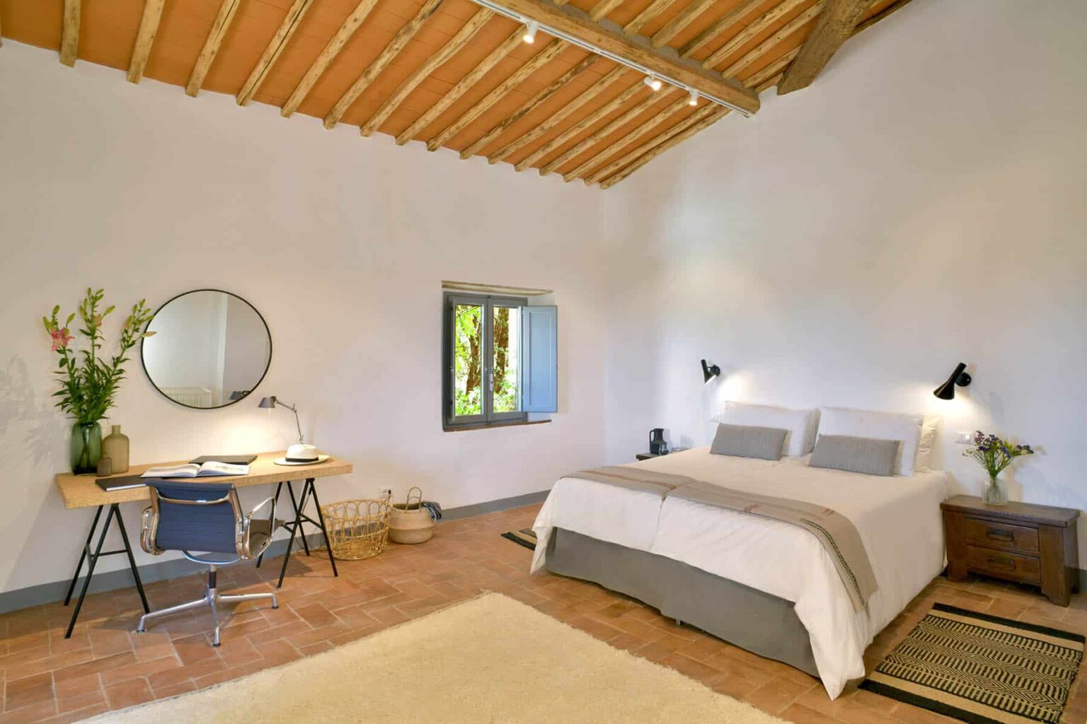 Spacious and modern bedroom featuring a queen-sized bed with neutral bedding, a stylish desk with a chair, and a round mirror. Natural light streams in through a window, highlighting the rustic wooden ceiling and minimalist decor. Ideal for relaxation and productivity. | Only on SopranoVillas