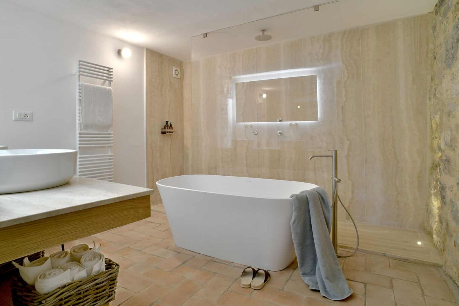Modern bathroom featuring a freestanding white bathtub, wooden vanity, and neutral stone walls. Soft lighting enhances the serene atmosphere, complemented by a towel draped over the tub and a basket for storage. | Only on SopranoVillas