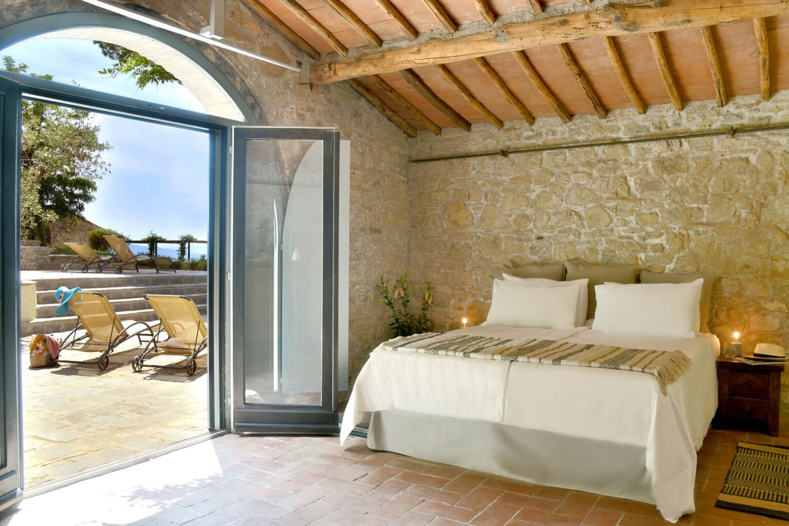 Cozy bedroom with a stone wall, featuring a comfortable double bed and large open doors leading to a sunlit terrace with loungers, ideal for relaxation and enjoying the outdoors. | Only on SopranoVillas