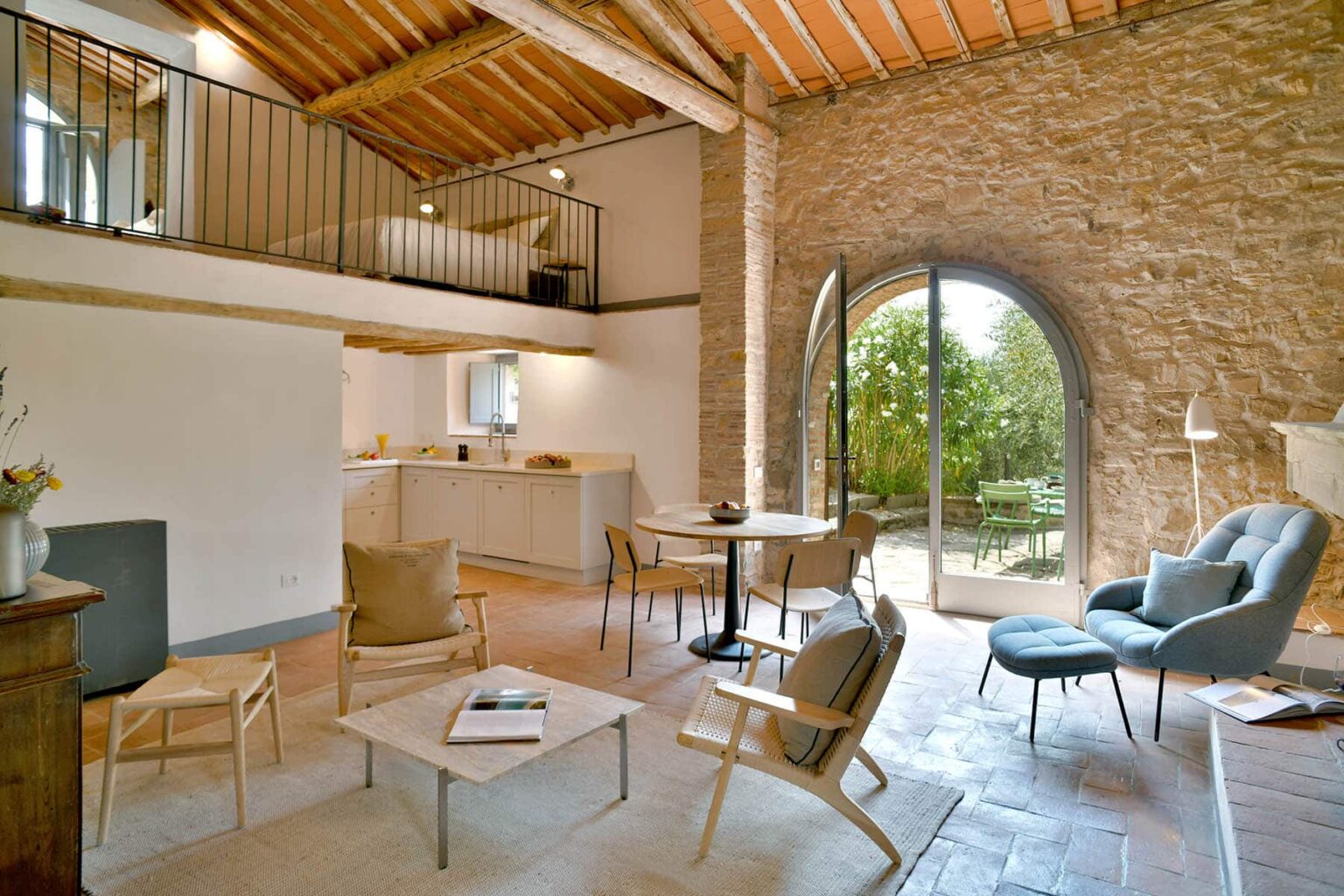 Modern living room with high wooden ceiling, natural stone walls, and large windows opening to a garden. The space features comfortable seating, a dining area, and a well-equipped kitchen, creating a bright and inviting atmosphere. | Only on SopranoVillas