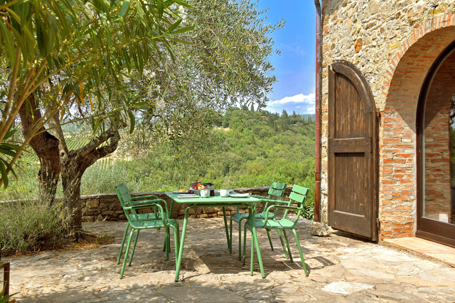 Outdoor dining area with green chairs and a table under an olive tree, showcasing a stone building and lush hillside in a tranquil setting. | Only on SopranoVillas