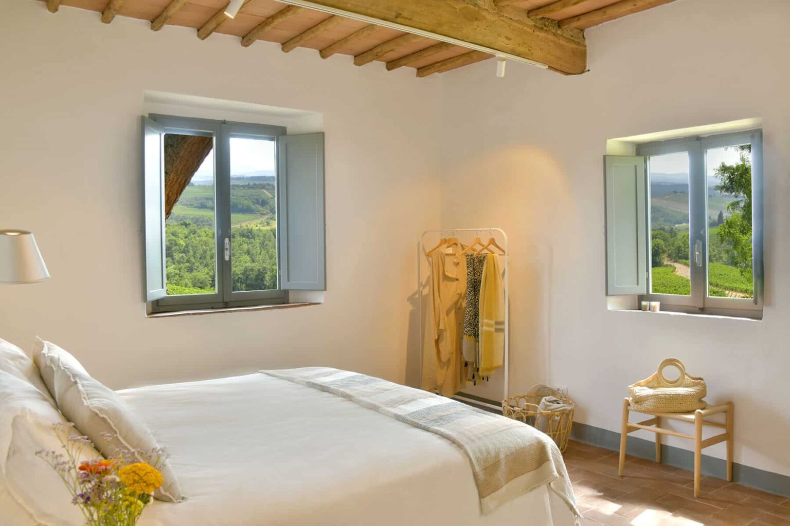 Bright, airy bedroom featuring a queen-size bed with neutral bedding, wooden beams on the ceiling, and large windows offering scenic views of greenery. A cozy chair and a decorative rack add to the inviting atmosphere. | Only on SopranoVillas