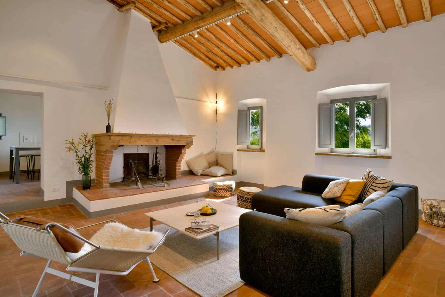Modern living room featuring a black sofa, a wooden fireplace, and exposed beam ceiling, with large windows allowing natural light and views of the outdoors. | Only on SopranoVillas
