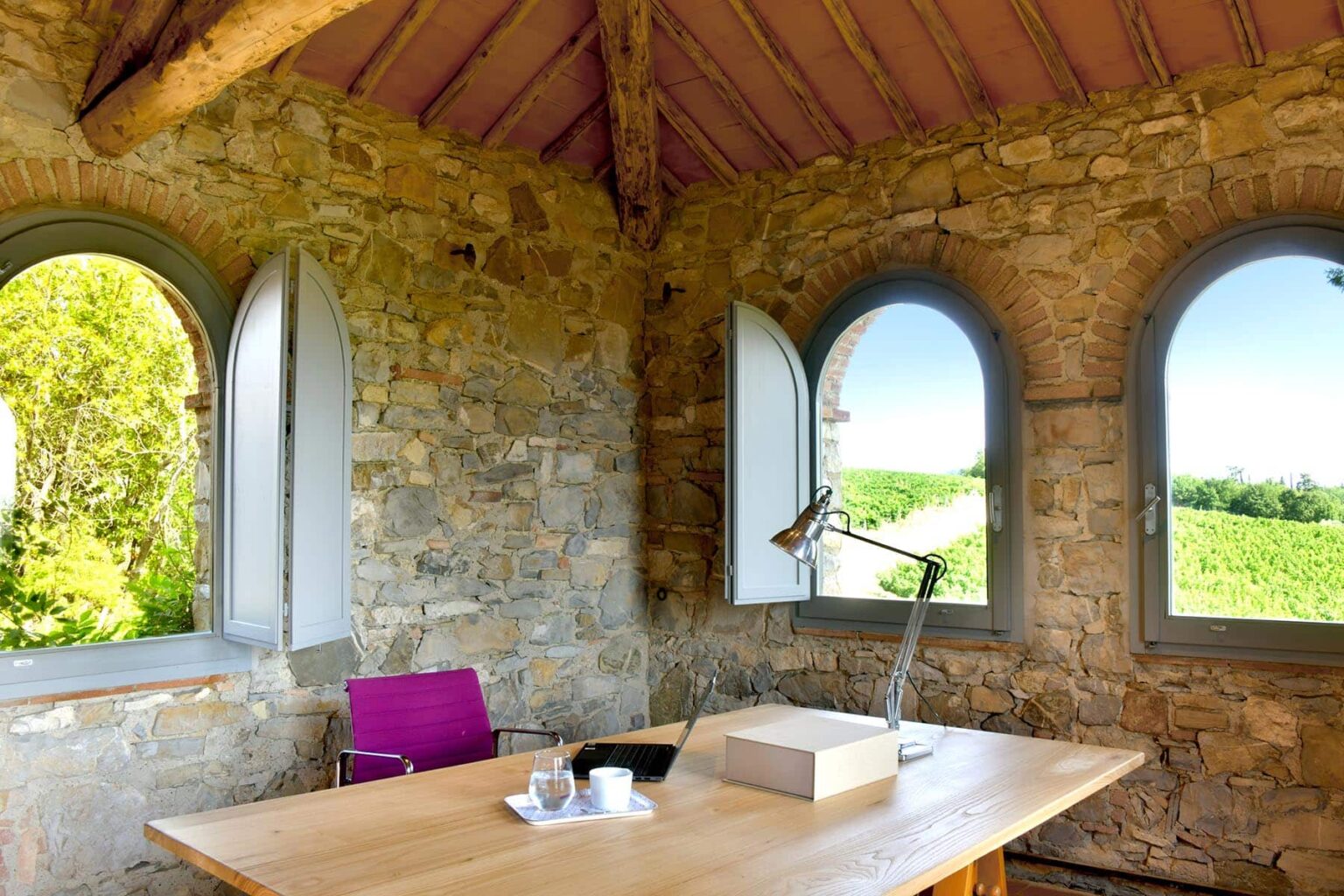 Cozy home office with stone walls and large arched windows overlooking lush greenery, featuring a wooden desk, a modern lamp, and a cup of tea. Ideal for remote work or creative inspiration in a serene setting. | Only on SopranoVillas