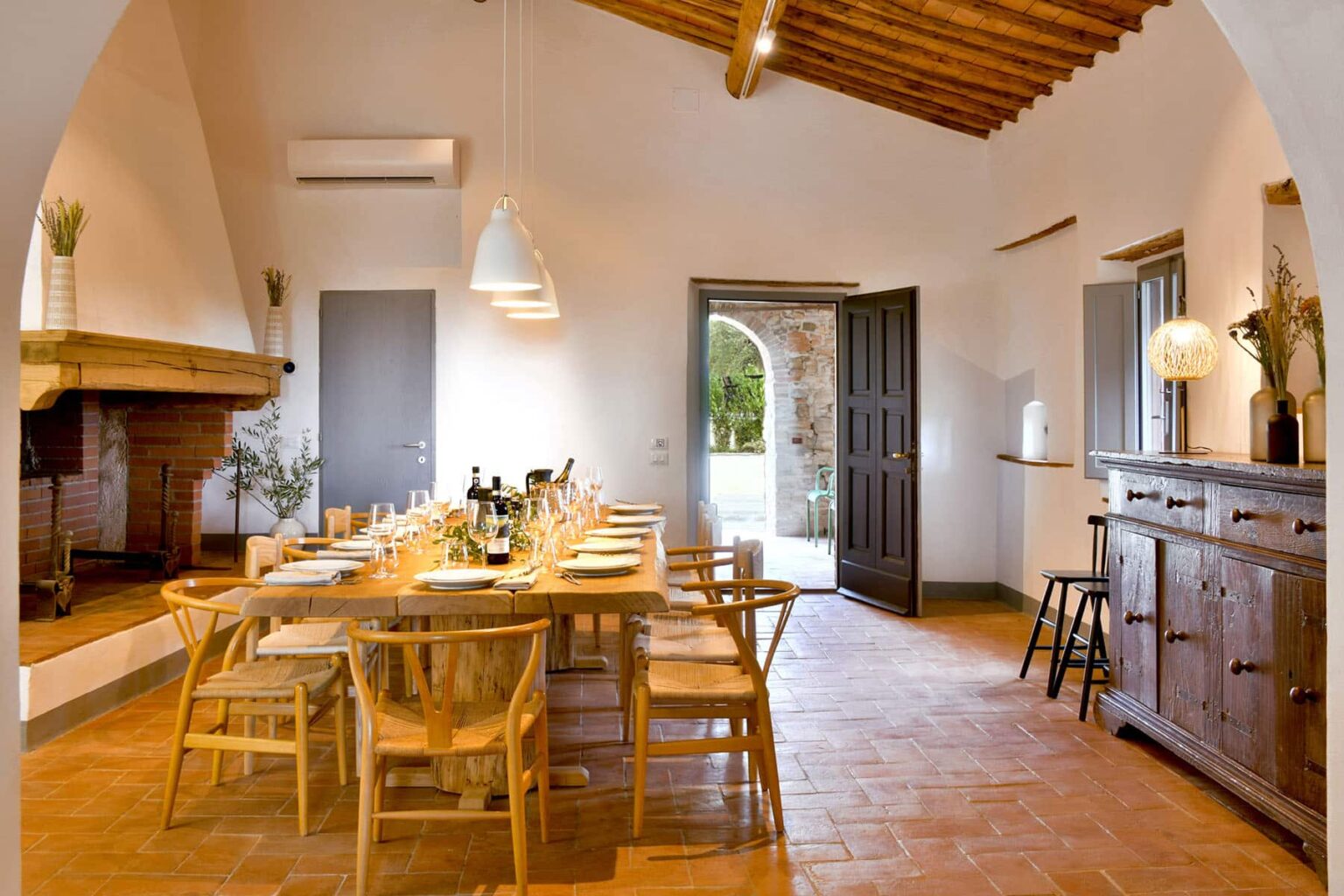 Modern dining room with a long wooden table set for a meal, featuring natural light from large doors, wooden beams on the ceiling, and minimalist decor. | Only on SopranoVillas
