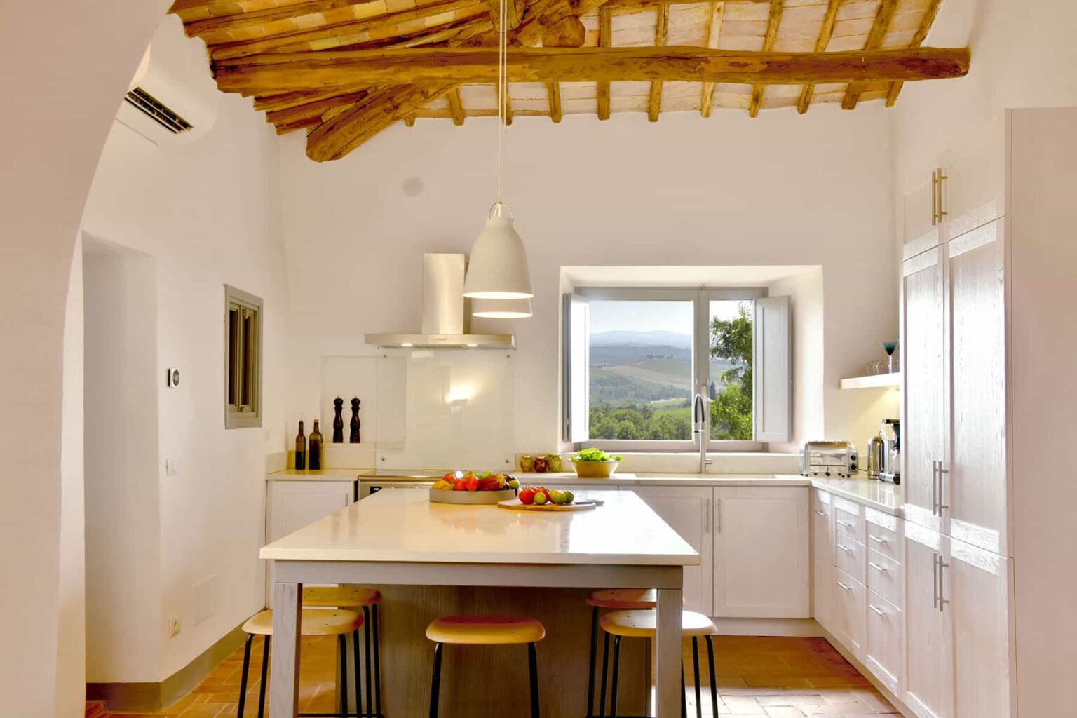 Modern kitchen with wooden ceiling beams, white cabinetry, and a central island featuring fresh produce. Large window offers a scenic view, enhancing the bright and airy atmosphere. | Only on SopranoVillas