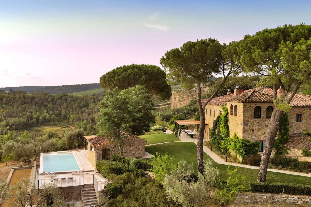 Scenic view of a rustic villa surrounded by lush greenery and tall trees, featuring a swimming pool and well-maintained gardens, set against a serene landscape in the countryside. | Only on SopranoVillas