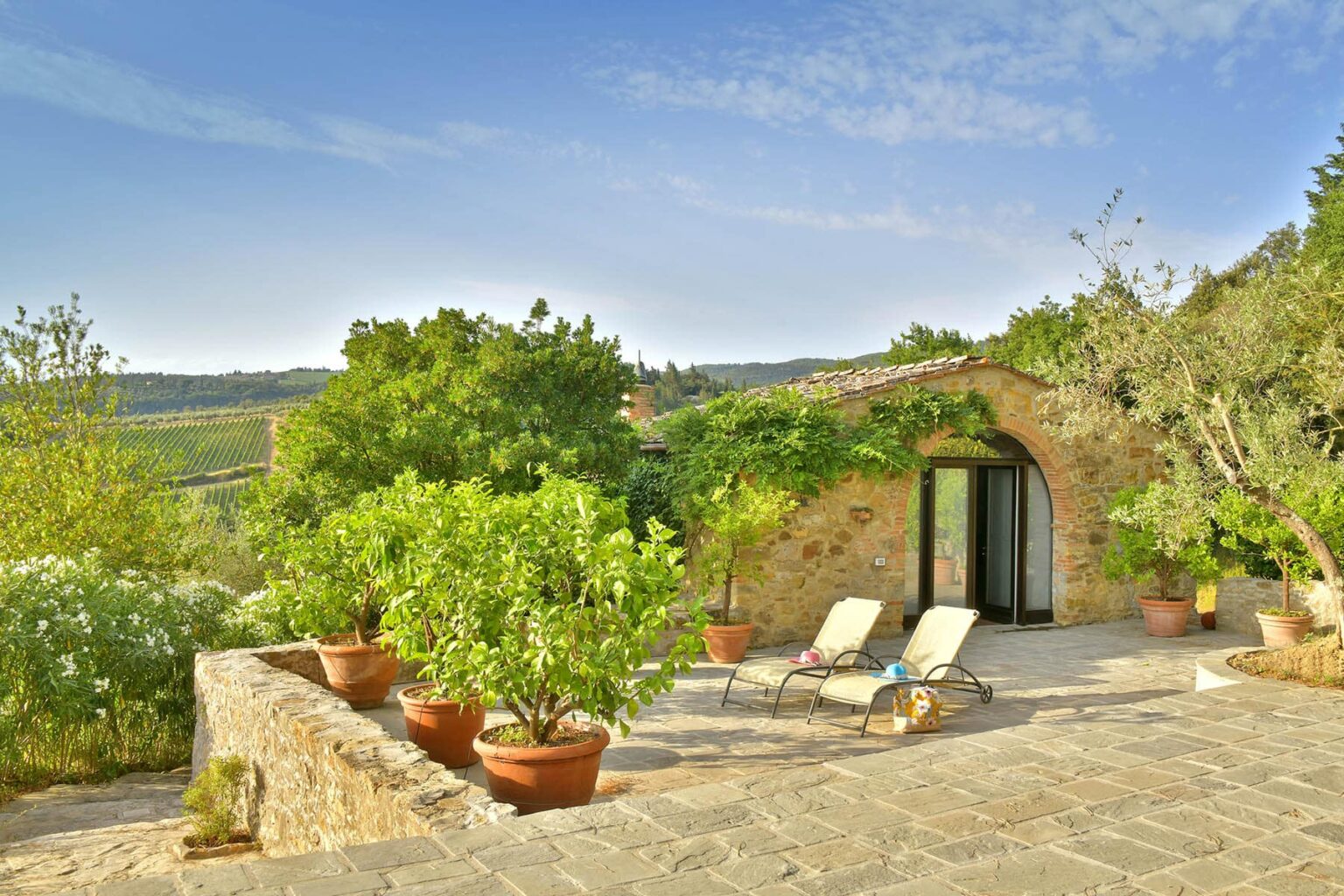 Charming rustic villa with a stone exterior, surrounded by lush greenery and potted plants, featuring two sun loungers on a spacious terrace under a clear blue sky. Ideal for relaxation in a serene countryside setting. | Only on SopranoVillas