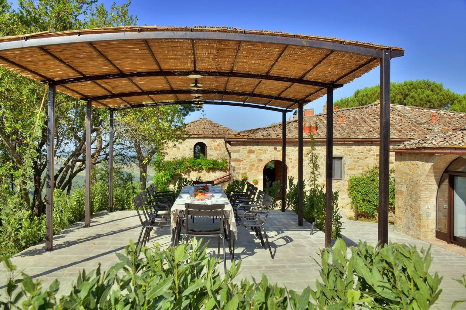 Outdoor dining area with a large table under a thatched roof, surrounded by lush greenery and a rustic stone building in the background, ideal for al fresco meals in a tranquil setting. | Only on SopranoVillas