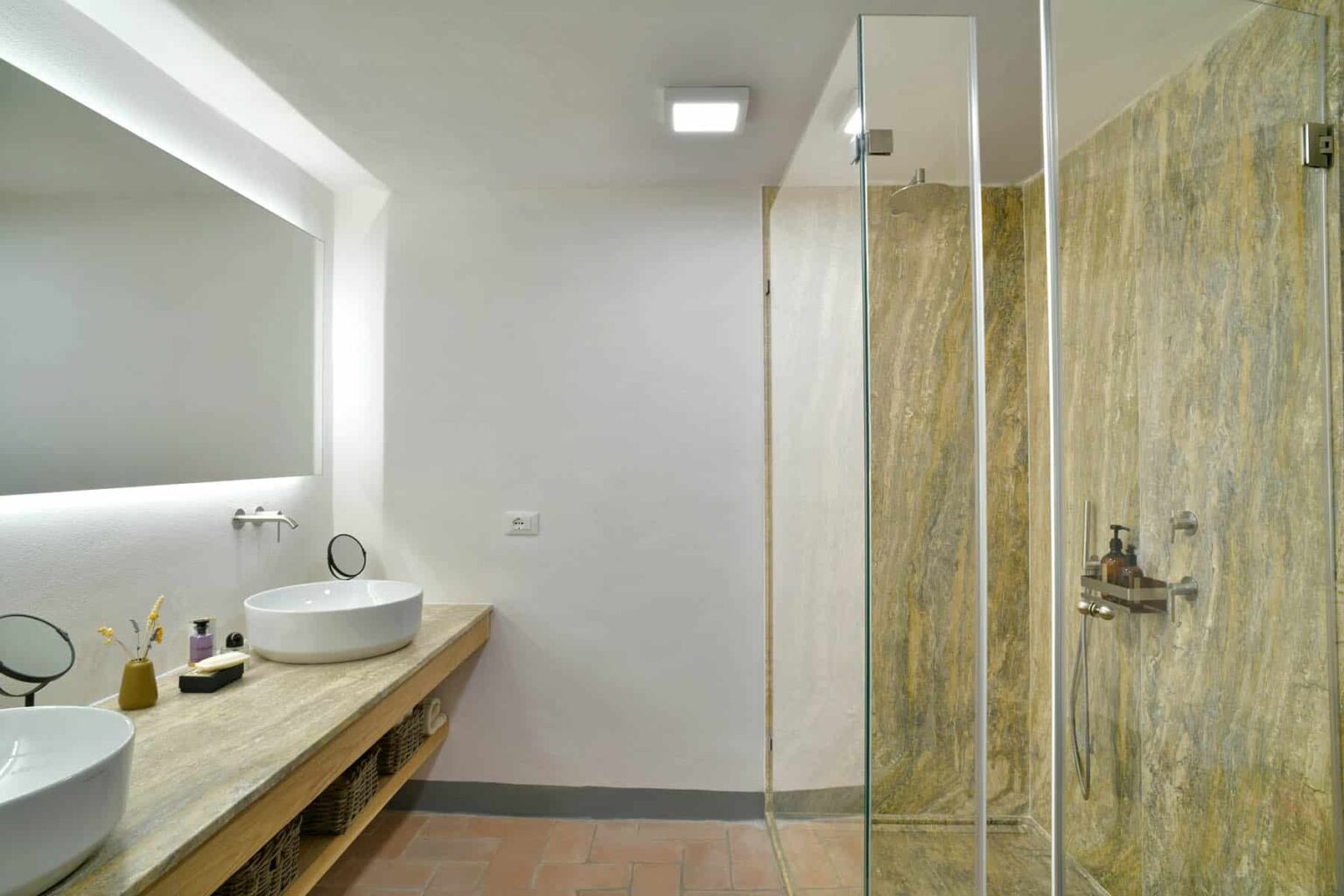 Modern bathroom featuring a glass shower enclosure, elegant stone wall, wooden vanity with round sinks, and stylish lighting for a contemporary aesthetic. | Only on SopranoVillas
