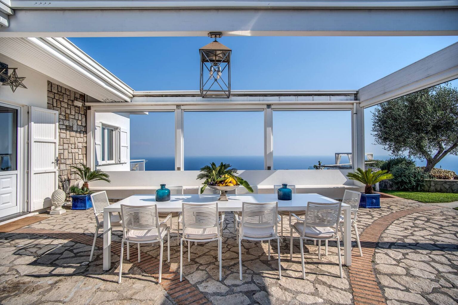 Outdoor dining area with a large white table and chairs, surrounded by lush greenery and a stunning ocean view under a pergola. Ideal for entertaining or relaxing in a scenic coastal setting. | Only on SopranoVillas