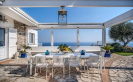 Outdoor dining area with a large white table and chairs, surrounded by lush greenery and a stunning ocean view under a pergola. Ideal for entertaining or relaxing in a scenic coastal setting.