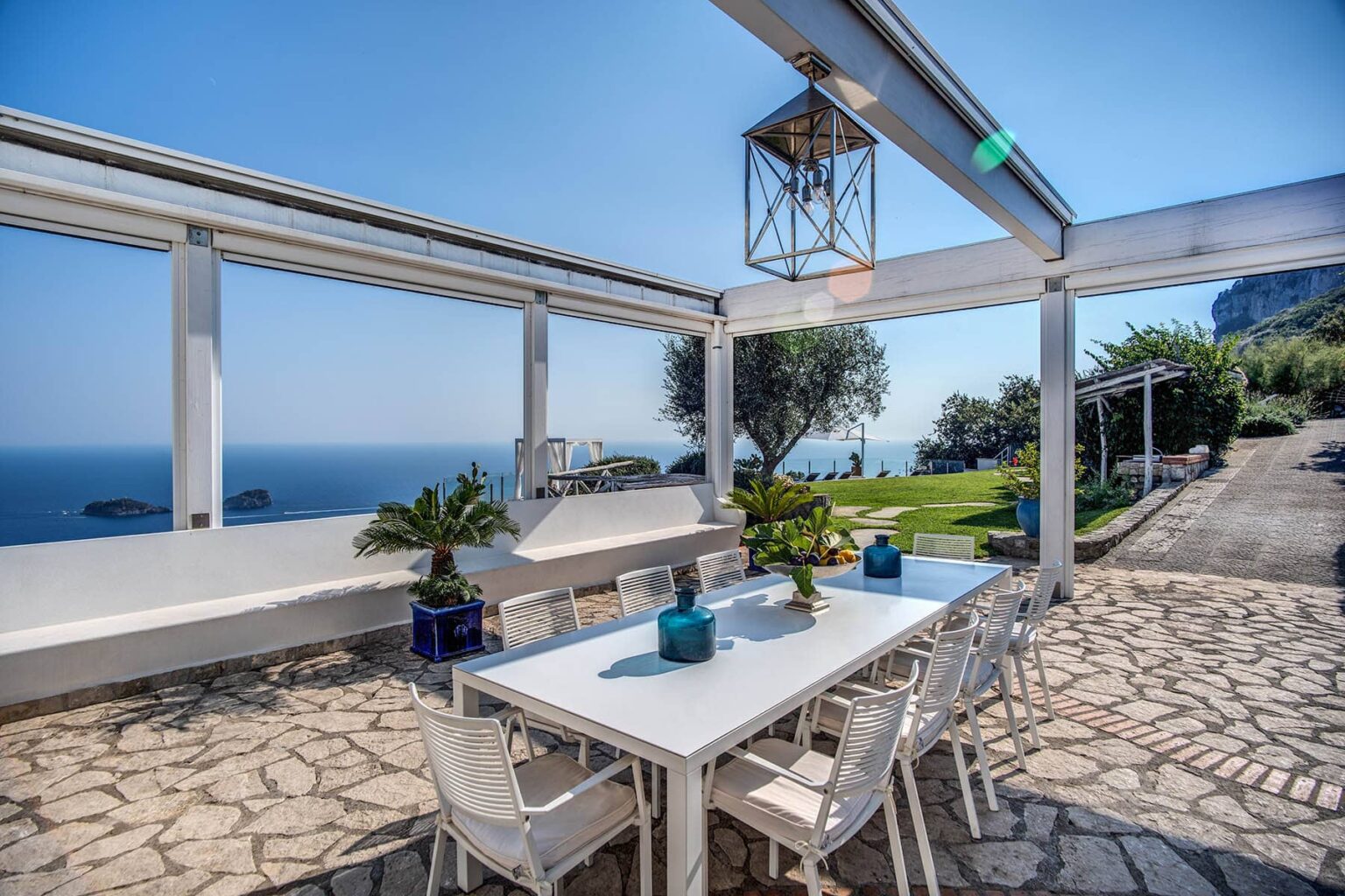 Outdoor dining area with a white table and chairs, overlooking a scenic ocean view, surrounded by greenery and a stone pathway, perfect for al fresco meals. | Only on SopranoVillas