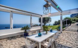 Outdoor dining area with a white table and chairs, overlooking a scenic ocean view, surrounded by greenery and a stone pathway, perfect for al fresco meals.