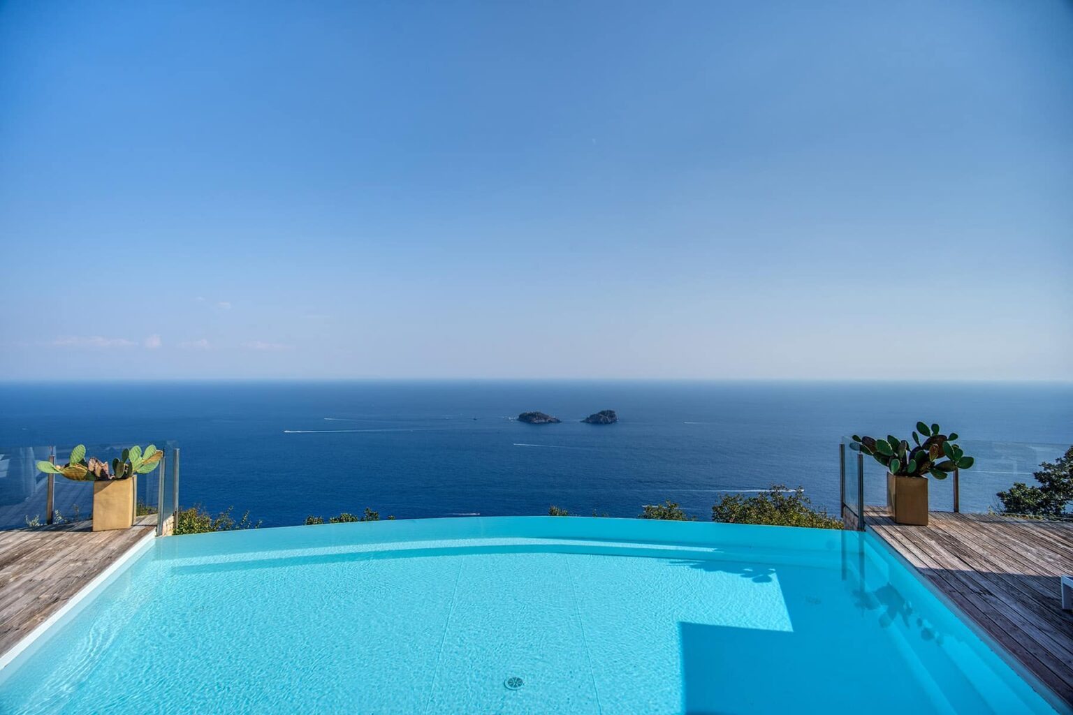 Infinity pool overlooking the ocean with clear blue water and distant islands on the horizon, surrounded by tropical plants and a wooden deck. Perfect for luxury vacation escapes and stunning coastal views. | Only on SopranoVillas