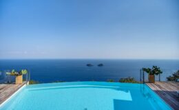 Infinity pool overlooking the ocean with clear blue water and distant islands on the horizon, surrounded by tropical plants and a wooden deck. Perfect for luxury vacation escapes and stunning coastal views.