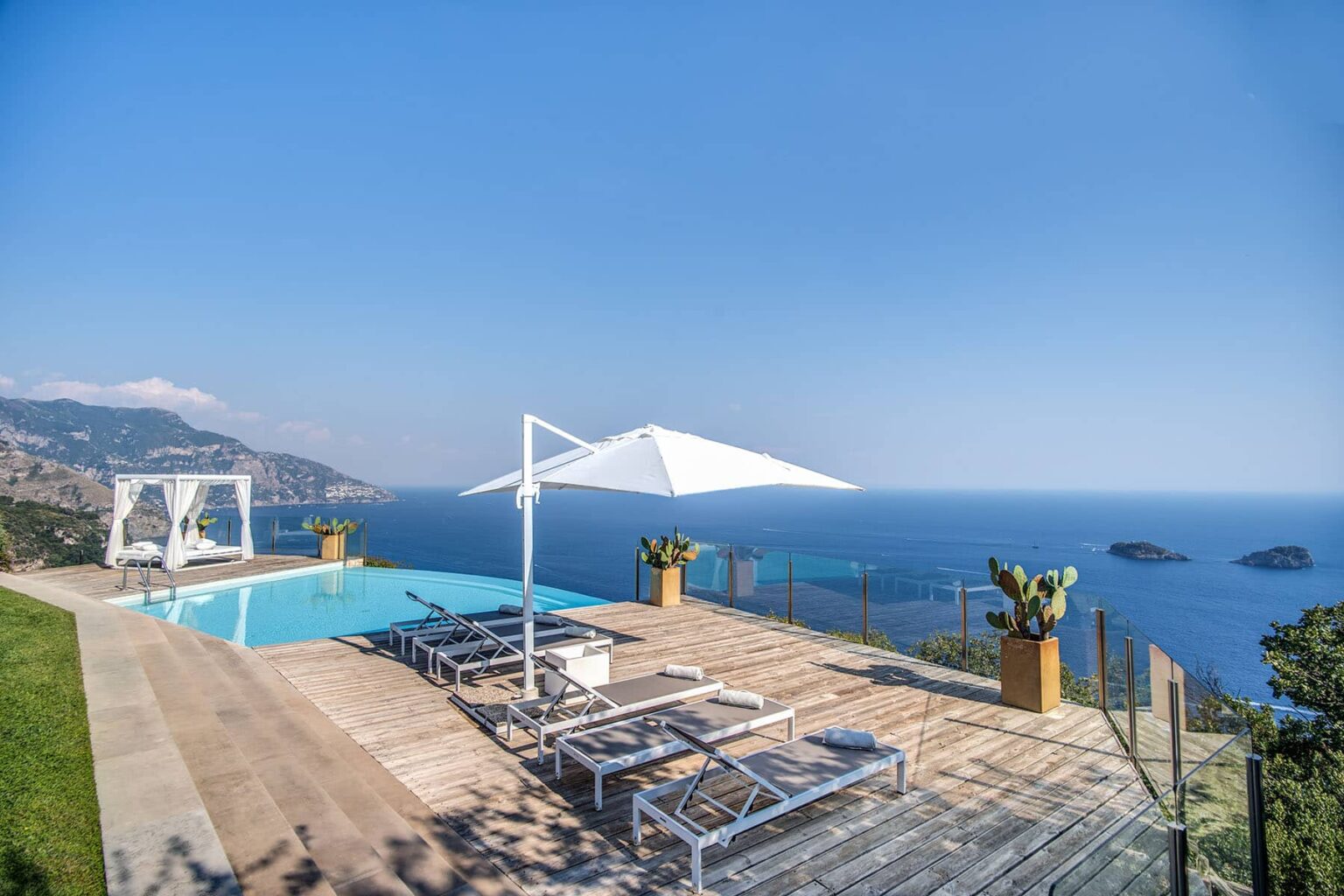 Luxury villa with a swimming pool overlooking the ocean, featuring sun loungers and a large umbrella, surrounded by lush greenery and scenic coastal views. | Only on SopranoVillas