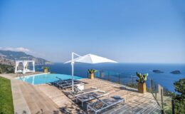 Luxury villa with a swimming pool overlooking the ocean, featuring sun loungers and a large umbrella, surrounded by lush greenery and scenic coastal views.