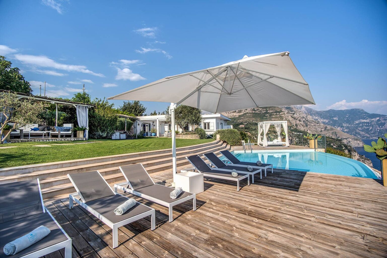 Modern outdoor pool area with lounge chairs and a large umbrella, surrounded by lush greenery and scenic mountain views, ideal for relaxation and leisure. | Only on SopranoVillas